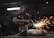 A worker in a dimly lit workshop is focused on welding two metal pieces together, causing bright sparks to fly. The workspace is cluttered with tools and materials, reflecting an industrial setting.