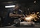 A worker in a dimly lit workshop is focused on welding two metal pieces together, causing bright sparks to fly. The workspace is cluttered with tools and materials, reflecting an industrial setting.