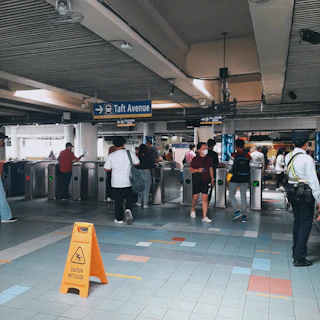 A busy subway or train station with multiple people walking through turnstiles. There is a sign indicating 'Taft Avenue' with directional arrows. A security guard is standing near a wet floor caution sign placed on the tiled floor. Several individuals are observed, some wearing face masks.