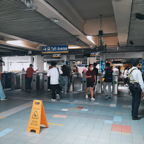 A busy subway or train station with multiple people walking through turnstiles. There is a sign indicating 'Taft Avenue' with directional arrows. A security guard is standing near a wet floor caution sign placed on the tiled floor. Several individuals are observed, some wearing face masks.