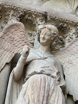 A stone sculpture of an angel with intricate details such as feathered wings and curly hair. The angel is holding a part of its garment, and the background features detailed carvings of leaves and vines.