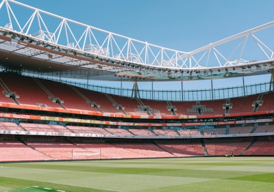 A large open-air football stadium with red seating and an expansive green field. The stadium is empty with sunlight streaming in, illuminating the seats and the pitch. The roof has a modern design with metal trusses and there are several advertising banners along the stands.