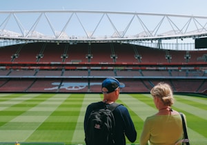 A large stadium with a vast green soccer field and surrounding red seats. Two people are standing in the foreground, looking over the stadium. The stadium's structure has a modern design with visible metal beams and open sky above.