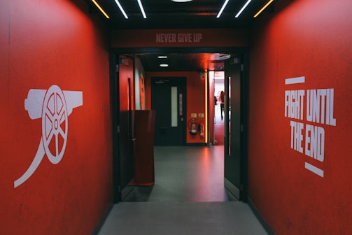 A hallway with vibrant red walls features motivational phrases such as 'Never Give Up' and 'Fight Until The End' in bold white lettering. On the left wall, there is an illustration of a cannon, symbolizing strength and resilience. The ceiling is adorned with several lines of light, creating an energetic atmosphere. At the end of the hallway, there is a doorway leading to a brighter area.