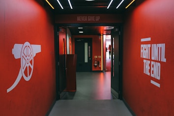 A hallway with vibrant red walls features motivational phrases such as 'Never Give Up' and 'Fight Until The End' in bold white lettering. On the left wall, there is an illustration of a cannon, symbolizing strength and resilience. The ceiling is adorned with several lines of light, creating an energetic atmosphere. At the end of the hallway, there is a doorway leading to a brighter area.