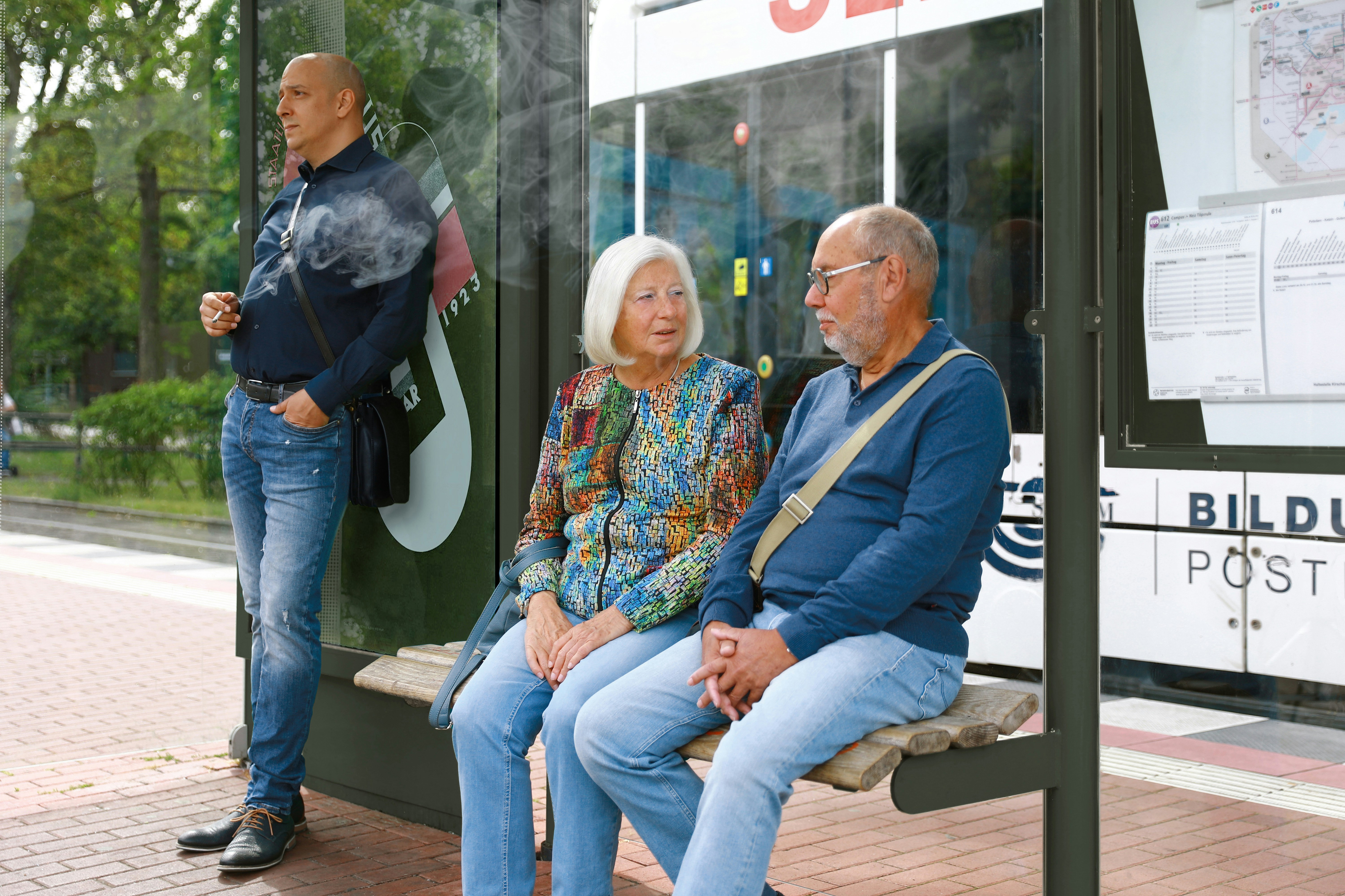A man waiting at a bus stop smokes a cigarette not aware that the smoke disturbs an elderly couple sitting at a bench - an appeal for a little more attention and awareness when smoking outdoors. Ein Mann raucht eine Zigarette während er auf die Tram wartet und bemerkt nicht, dass der Rauch ein älteres Pärchen stört - ein Aufruf zu etwas mehr Rücksicht und Achtsamkeit beim Rauchen im Freien.