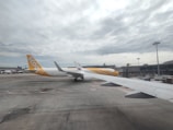 A large passenger airplane from Scoot Airlines is stationed on the tarmac at an airport. The aircraft is predominantly white with distinctive yellow accents, including the tail fin. The sky is cloudy, and other airport infrastructure, such as terminal buildings and ramps, can be seen in the background.