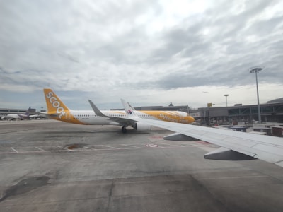 A large passenger airplane from Scoot Airlines is stationed on the tarmac at an airport. The aircraft is predominantly white with distinctive yellow accents, including the tail fin. The sky is cloudy, and other airport infrastructure, such as terminal buildings and ramps, can be seen in the background.