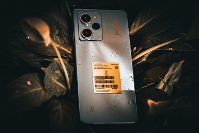 A smartphone with multiple rear cameras is placed on leaves, with water droplets visible on its surface. The phone has a label attached, detailing specifications and manufacturing information.