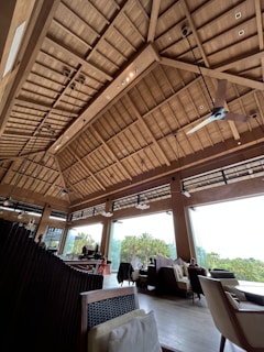 A spacious interior with a high wooden ceiling featuring beams and modern light fixtures. Large windows allow natural light to flood the area, providing a view of lush greenery outside. Comfortable seating with cushions is arranged throughout the space. Pendant lights and a ceiling fan add to the contemporary decor.
