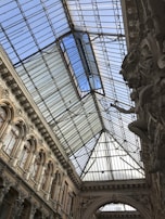 An intricately designed architectural interior featuring a large glass ceiling with steel framework. The walls display detailed carvings and ornate arches with series of windows along the structure, showcasing classic European design elements.