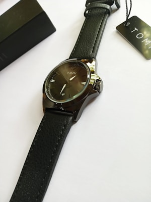 A sleek wristwatch with a black leather strap and a black dial. The watch face features minimalistic markers and is branded with the name 'TOMI'. It is placed on a white surface with a black box and a tag nearby.