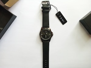 A black leather-strap wristwatch with a minimalist design and the brand name Tomi on the dial. The watch is neatly placed on a plain white background with two black watch boxes and a tag labeled Tomi nearby.