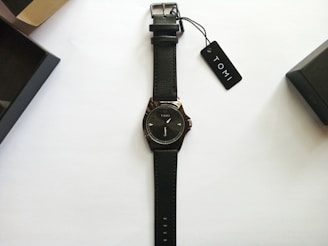 A black leather-strap wristwatch with a minimalist design and the brand name Tomi on the dial. The watch is neatly placed on a plain white background with two black watch boxes and a tag labeled Tomi nearby.