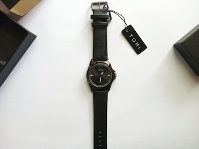A black leather-strap wristwatch with a minimalist design and the brand name Tomi on the dial. The watch is neatly placed on a plain white background with two black watch boxes and a tag labeled Tomi nearby.