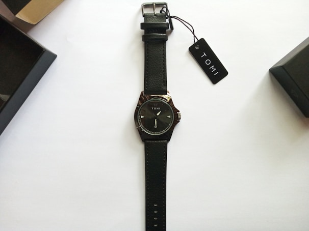 A black leather-strap wristwatch with a minimalist design and the brand name Tomi on the dial. The watch is neatly placed on a plain white background with two black watch boxes and a tag labeled Tomi nearby.