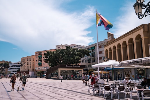 A lively town square surrounded by buildings featuring varied architectural styles, with a prominent flagpole displaying a colorful flag. People are leisurely strolling or sitting at outdoor café tables enjoying the sunny day. A large tree provides shade, and the presence of a lamppost and a clock enhance the urban setting.
