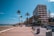 A beachfront promenade flanked by tall palm trees and modern apartment buildings. The foreground features a path lined with benches and trash bins. To the left, a sandy beach extends into the distance, with the ocean visible under a clear blue sky. Parked cars line the street adjacent to the buildings.