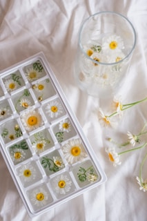An ice tray filled with ice cubes containing chamomile flowers and green leaves is placed on a white fabric surface. Next to the ice tray, there is a glass filled with similar floral ice cubes. A few loose chamomile flowers are scattered around the glass.