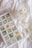 An ice tray filled with ice cubes containing chamomile flowers and green leaves is placed on a white fabric surface. Next to the ice tray, there is a glass filled with similar floral ice cubes. A few loose chamomile flowers are scattered around the glass.