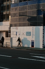 Two construction workers wearing safety helmets are walking near a construction site under warm, late afternoon light. The building exterior is covered with signage related to sustainable development goals and a temporary uniform wall enclosing the site.