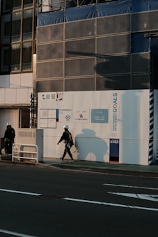 Two construction workers wearing safety helmets are walking near a construction site under warm, late afternoon light. The building exterior is covered with signage related to sustainable development goals and a temporary uniform wall enclosing the site.