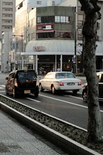 An urban street scene featuring several cars driving along a road bordered by a sidewalk. There's a large building in the background with a distinctive green exterior and a billboard displaying a wine bottle. The area appears to be in a busy city setting, with traffic signs and a tree in the foreground.
