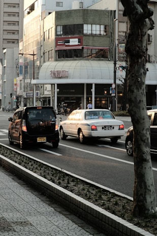 An urban street scene featuring several cars driving along a road bordered by a sidewalk. There's a large building in the background with a distinctive green exterior and a billboard displaying a wine bottle. The area appears to be in a busy city setting, with traffic signs and a tree in the foreground.