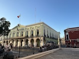 A large historic building with neoclassical architecture is prominent, showcasing green and white walls with tall arched windows and ornate details. A Mexican flag is flying on top of the building. In the foreground, people are walking, and there is a black wrought iron fence bordering the area. The sky is clear and blue, suggesting a sunny day.