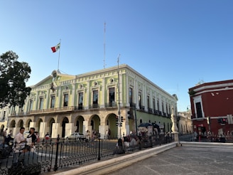 A large historic building with neoclassical architecture is prominent, showcasing green and white walls with tall arched windows and ornate details. A Mexican flag is flying on top of the building. In the foreground, people are walking, and there is a black wrought iron fence bordering the area. The sky is clear and blue, suggesting a sunny day.