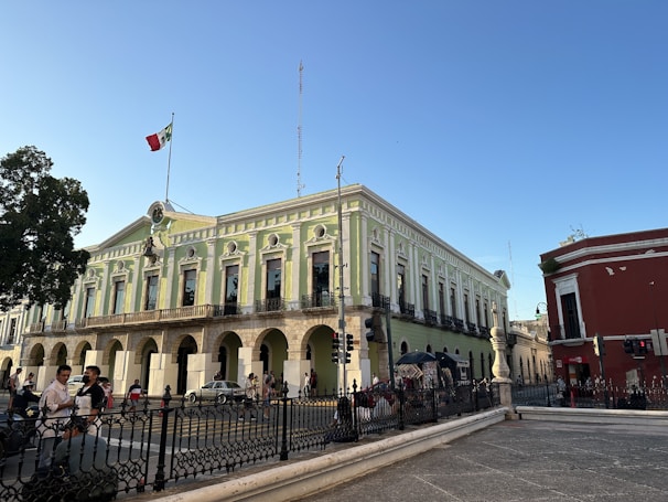 A large historic building with neoclassical architecture is prominent, showcasing green and white walls with tall arched windows and ornate details. A Mexican flag is flying on top of the building. In the foreground, people are walking, and there is a black wrought iron fence bordering the area. The sky is clear and blue, suggesting a sunny day.