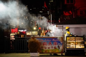 Two street vendors appear to be working at a food stall at night. One vendor is wearing a yellow helmet and jacket, possibly a delivery uniform, while preparing food. The other vendor, in dark clothing, seems to be cooking, with visible steam rising. The scene is set against the backdrop of an urban environment with illuminated signage and dimly lit buildings.