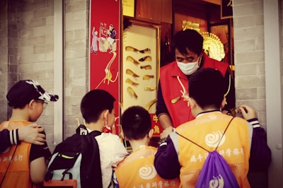 A group of children wearing orange vests and backpacks are gathered around a person in a red vest and mask, standing in front of a shop with traditional decorations. The setting suggests a lively street or market.