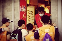 A group of children wearing orange vests and backpacks are gathered around a person in a red vest and mask, standing in front of a shop with traditional decorations. The setting suggests a lively street or market.