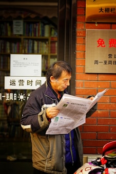 An older man reads a newspaper outdoors near the entrance of a building with signs written in Chinese. The background includes a glass door showing books on shelves inside.