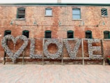 A brick wall with a large artistic installation featuring the word 'LOVE' created from numerous colorful locks. The installation includes a heart shape on the left and is set in an outdoor setting with a cobblestone ground. Green lamps are mounted on the wall, adding an industrial look.
