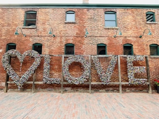 A brick wall with a large artistic installation featuring the word 'LOVE' created from numerous colorful locks. The installation includes a heart shape on the left and is set in an outdoor setting with a cobblestone ground. Green lamps are mounted on the wall, adding an industrial look.