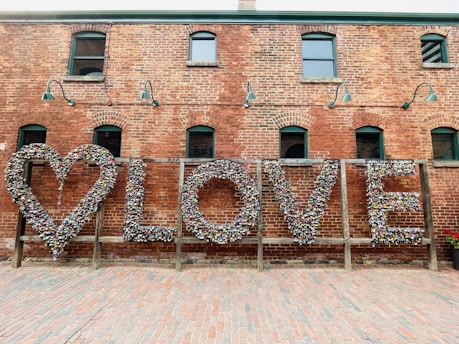 A brick wall with a large artistic installation featuring the word 'LOVE' created from numerous colorful locks. The installation includes a heart shape on the left and is set in an outdoor setting with a cobblestone ground. Green lamps are mounted on the wall, adding an industrial look.