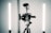 A professional video camera is mounted on a tripod with a microphone attached on top. Two vertical LED lights are positioned on either side of the camera for illumination. The setup appears to be in a studio environment with a minimalist background.