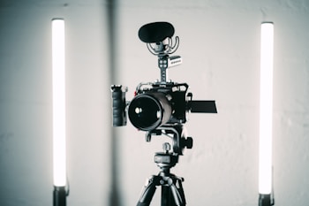 A professional video camera is mounted on a tripod with a microphone attached on top. Two vertical LED lights are positioned on either side of the camera for illumination. The setup appears to be in a studio environment with a minimalist background.