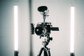 A professional video camera is mounted on a tripod with a microphone attached on top. Two vertical LED lights are positioned on either side of the camera for illumination. The setup appears to be in a studio environment with a minimalist background.