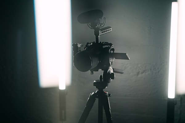 A professional video camera mounted on a tripod stands between two vertical LED lights in a dimly lit environment, giving the scene a cinematic look.
