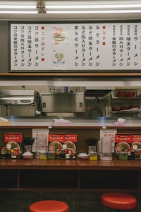 A cozy ramen shop with a wooden counter featuring red stools. The counter is adorned with menu items, condiments like soy sauce and vinegar, and utensils. Above the counter, a large menu board with various ramen options is displayed in Japanese.