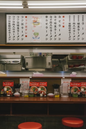 A cozy ramen shop with a wooden counter featuring red stools. The counter is adorned with menu items, condiments like soy sauce and vinegar, and utensils. Above the counter, a large menu board with various ramen options is displayed in Japanese.