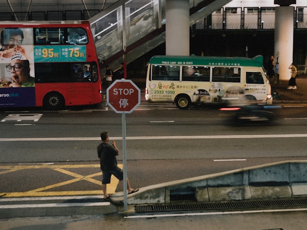 A red double-decker bus and a green minibus are parked on the side of a busy street, with a blurred motorbike passing by. A man stands at a crosswalk beside a stop sign featuring both English and Chinese characters. In the background, a pedestrian bridge and more people are visible along the sidewalk.