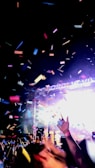 A lively concert scene featuring a crowd of people raising their hands amidst colorful confetti falling from above. Bright lights illuminate the stage in the background, creating a vibrant and energetic atmosphere.