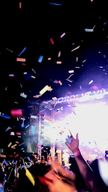 A lively concert scene featuring a crowd of people raising their hands amidst colorful confetti falling from above. Bright lights illuminate the stage in the background, creating a vibrant and energetic atmosphere.