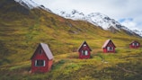 Four small red cabins with triangular roofs are scattered across a grassy hillside. The background features impressive snow-capped mountains under a cloudy sky, suggesting a remote, serene mountain setting.