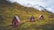 Four small red cabins with triangular roofs are scattered across a grassy hillside. The background features impressive snow-capped mountains under a cloudy sky, suggesting a remote, serene mountain setting.