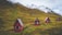 Four small red cabins with triangular roofs are scattered across a grassy hillside. The background features impressive snow-capped mountains under a cloudy sky, suggesting a remote, serene mountain setting.